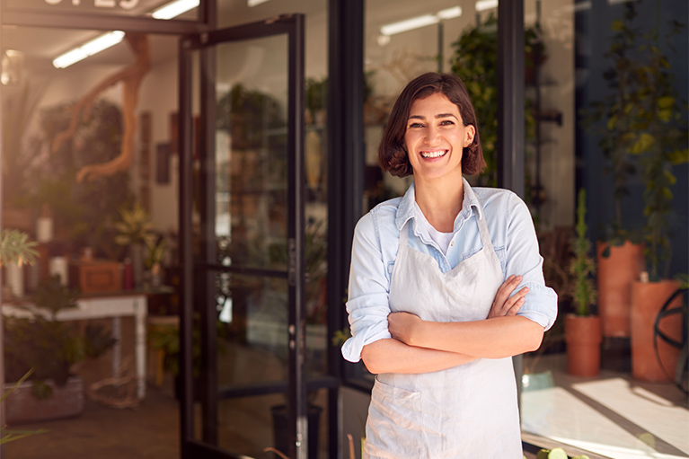 Smiling small business owner wearing an apron, standing confidently at the entrance of her shop.