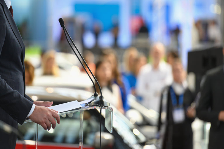 A person stands at a podium, holding papers, addressing an audience. The background is blurred, showing people standing and listening to a geopolitical webinar.