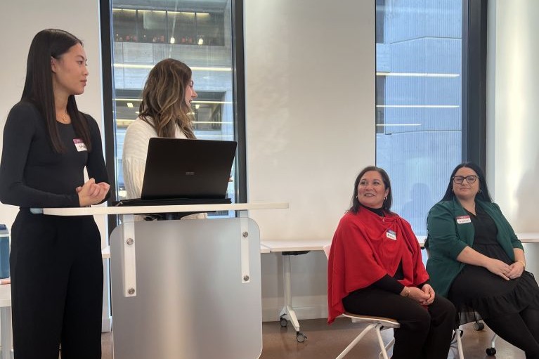 Two people standing at a podium with a laptop and two panelists sitting on chairs.