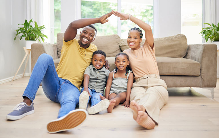 Parents sitting on the floor of their living room, making an arch with their arms over their two children who are sitting between them. 