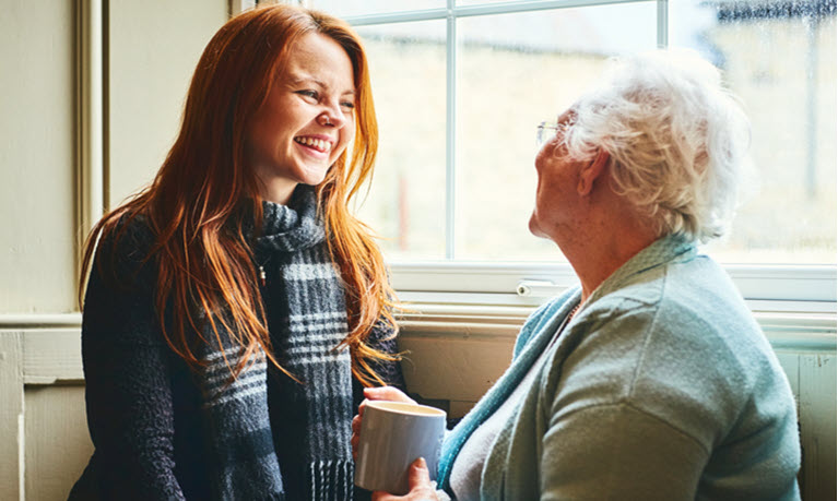 A woman with ginger hair smiling at an older woman who is holding a mug.