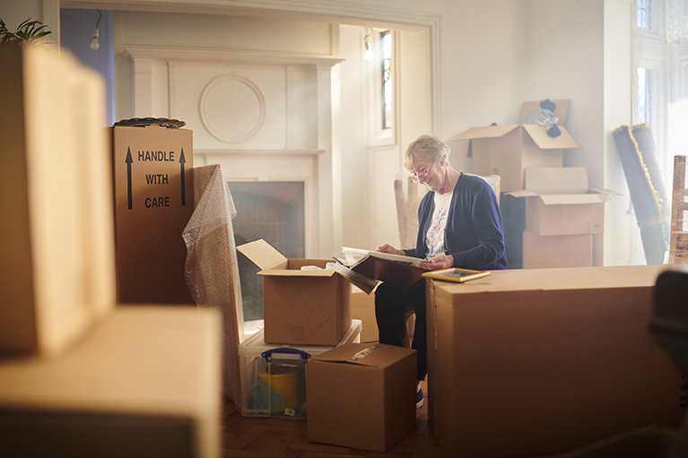 a senior sits among moving boxes looking at contents 
