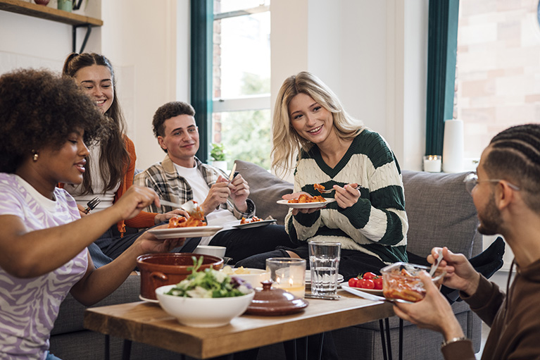 A low angle side view of group of friends who are students in the common room having a meal on the couch