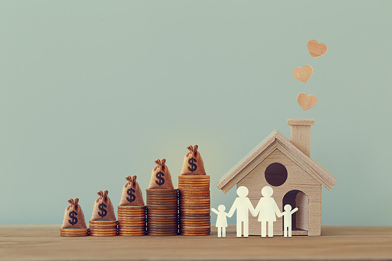 litte wooden house with family cut out of paper in front and coins and bags of money beside the house
