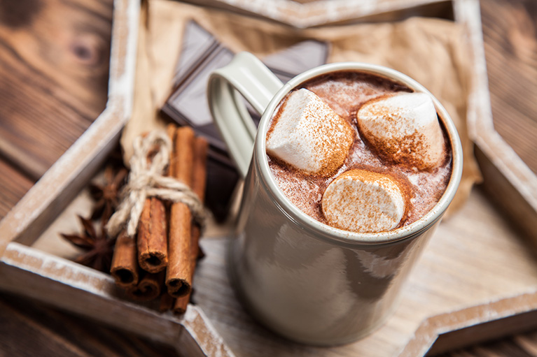 a wooden tray holding chocolate pieces, cinnamon sticks and a mug of hot chocolate with marshmallows