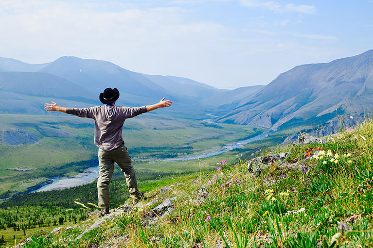man with hat standing with arms open wide on grassy hill overlooking hilly landscape