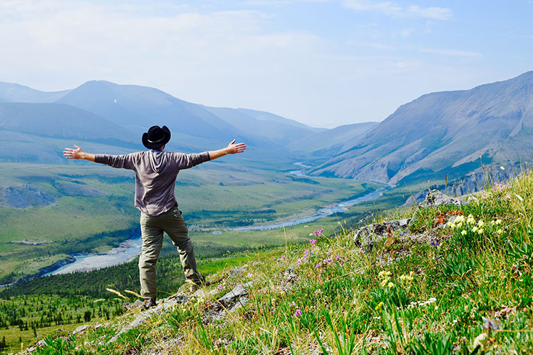 man with hat standing with arms open wide on grassy hill overlooking hilly landscape