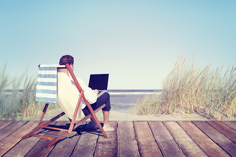 man sitting on a beach lounger on a wooden deck facing water with laptop on his lap