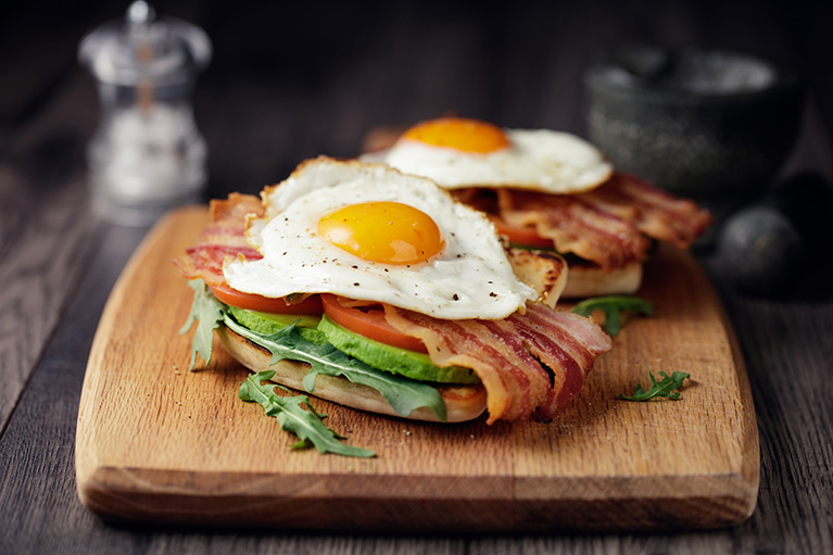 wooden board showing two open face sandwiches with avocado, tomato, bacon and sunnyside egg on top