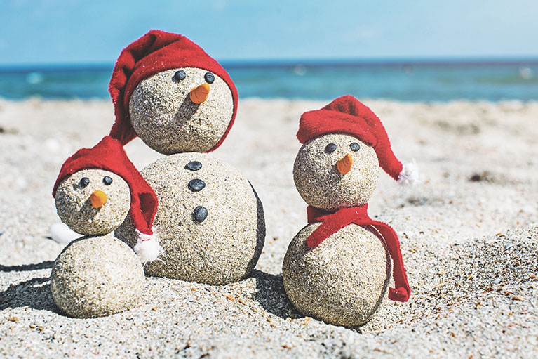 3 snowmen made of sand wearing red toques on a beach