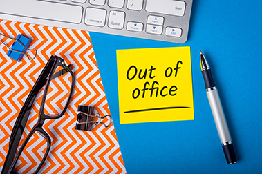 image showing part of computer keyboard, a pair of glasses and a sticky note with words "out of office" written on it