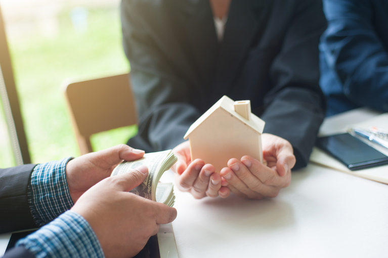 woman's hand holding a small wooden house and man's hands holding a stack of money at a table