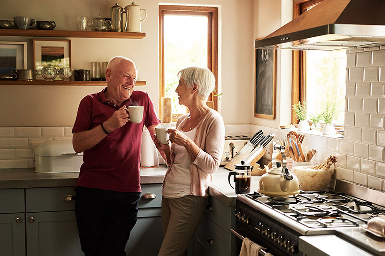 A man and a woman talking in the kitchen. Each of them have a cup in their hand.