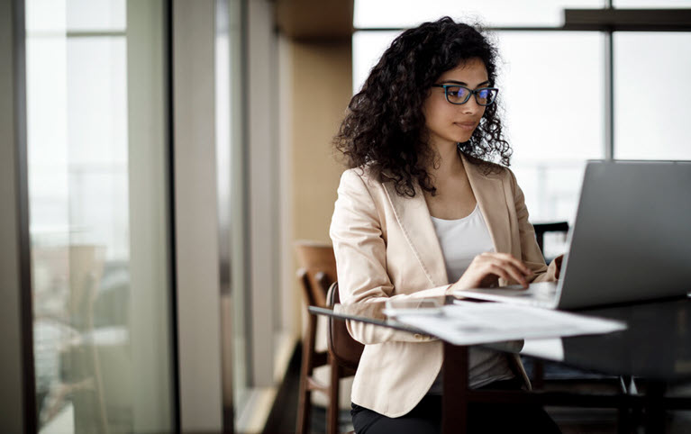 A woman sitting at a desk reading the National Bank Financial Markets' Forex monthly publication on her laptop. 