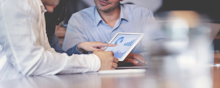 Three people sitting at a desk looking at a tablet with graphs on it.