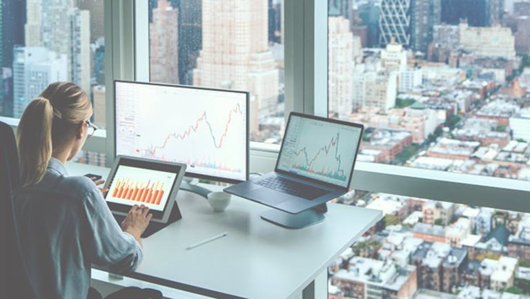 A woman sitting at a desk overlooking a city, working on a tablet with two other computers behind.
