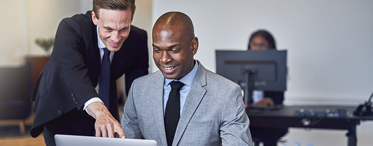 Two smiling men looking at a computer screen. One of them is pointing a finger at the screen.