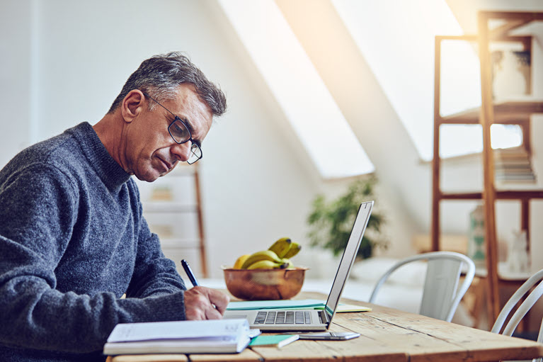 A man writing in a book at his desk with a laptop in front of him.