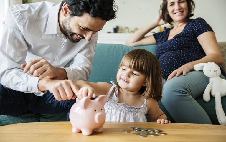 A little girl putting a coin into a piggy bank with her pregnant mother and father beside her.