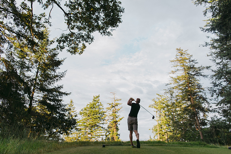 A man hits a ball on a forested golf course.
