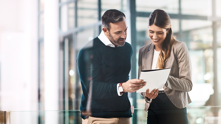 A man and a woman look at a notebook in a glass office.