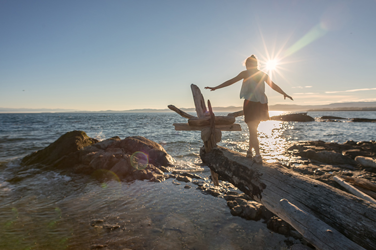 Lens flare on millenial young woman balancing on elevated log over rocky ocean beach at Holland Point Park, Victoria, British Columbia, Canada
