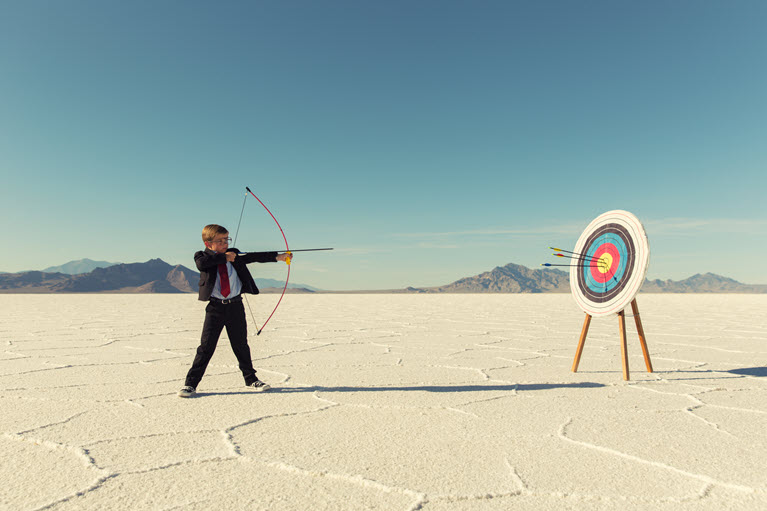 A boy with a bow firing an arrow at a target in the desert.