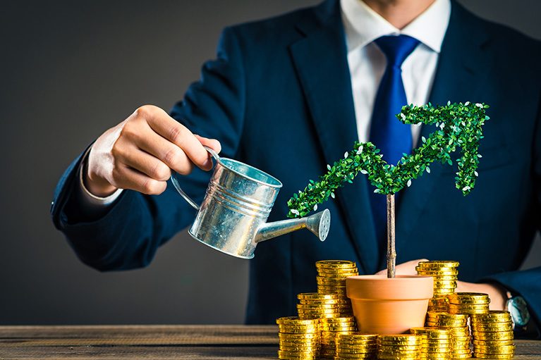 A plant surround by gold coins in front of a person in a suit with a watering can