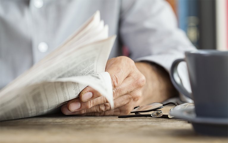 A man’s hand holding a newspaper at a desk which has a coffee mug  on it.