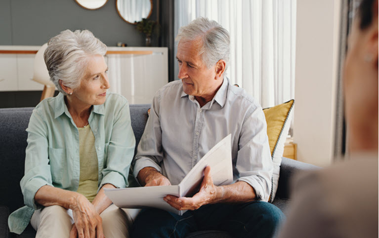 An elderly couple reviews documents on a couch with a third person, suggesting a discussion about estate planning.
