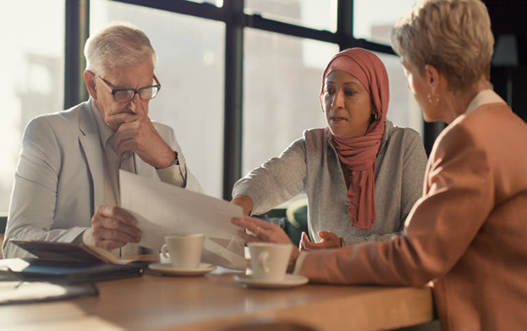 Three people discuss documents at a table in a bright room, symbolizing collaboration and review in portfolio management.
