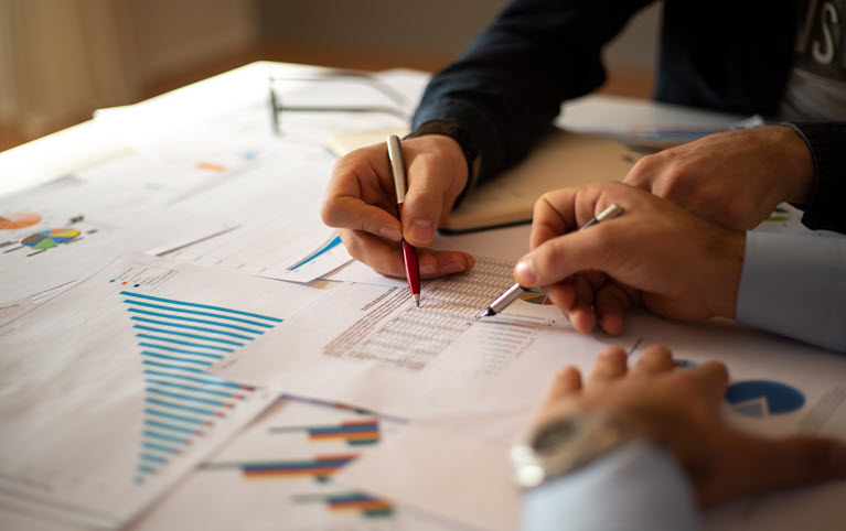 Two people review charts and graphs on a table, collaborating on financial planning with pens in hand.