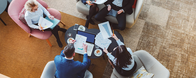 Five people in a business meeting, reviewing documents and charts around a coffee table.