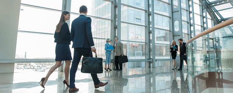 People walking in a modern glass building with large windows and reflections on the shiny floor. 