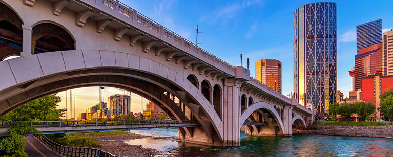 White arched bridge over a river with a city skyline in the background at sunset.